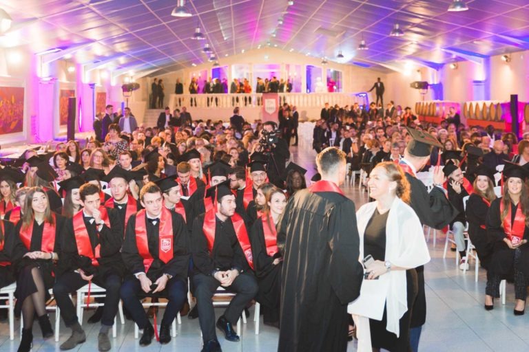 Une grande soirée de remise des diplômes de l'EBBS Bordeaux se déroule en intérieur et réunit les diplômés assis en rangs, vêtus de robes noires et de casquettes associées à des écharpes rouges. La première rangée met en valeur les diplômés attentifs, tandis que les intervenants et les officiels se tiennent au premier plan. La salle illuminée par des lumières bleues et violettes crée une atmosphère festive.