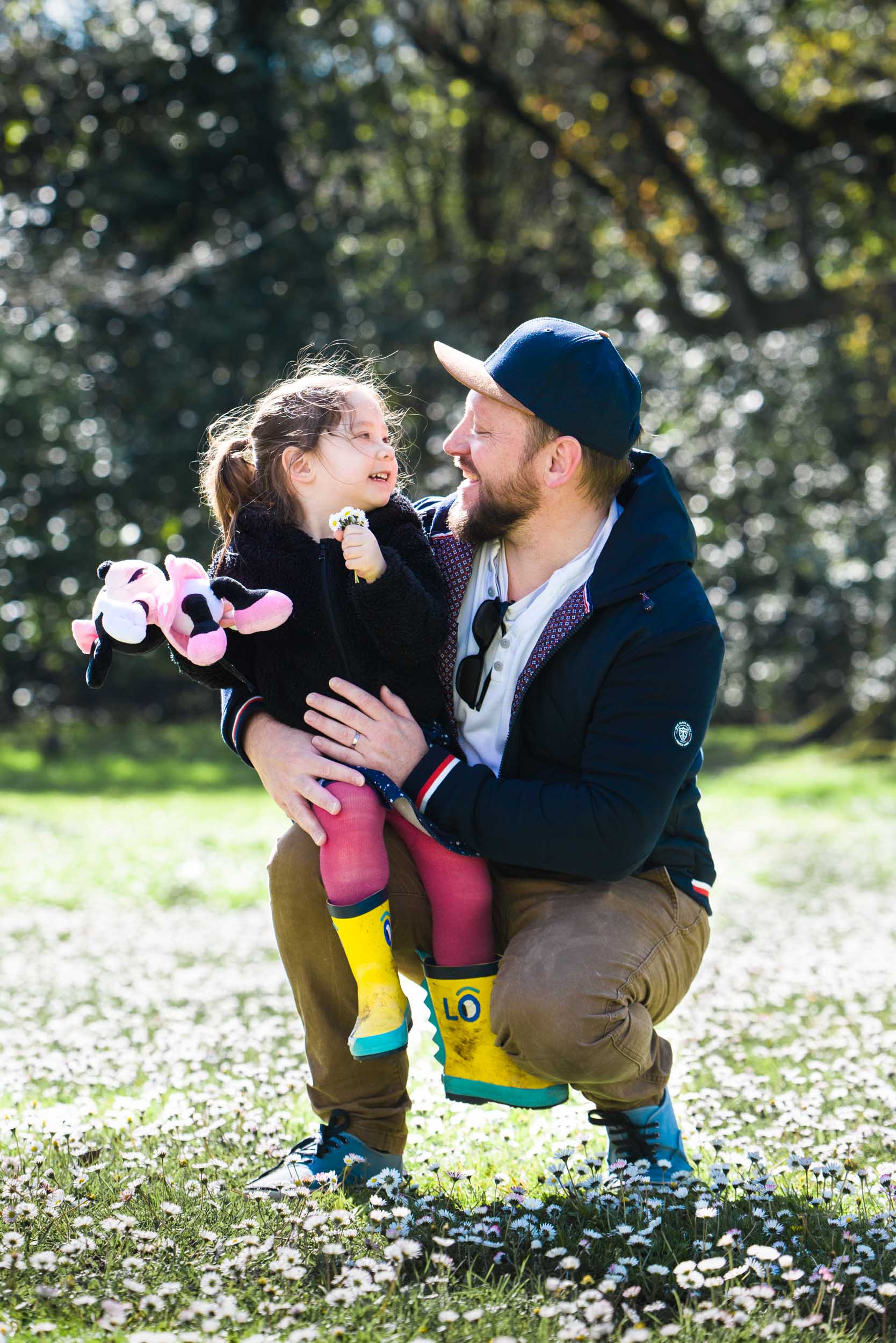 Un homme portant une casquette bleue et un sweat à capuche est accroupi sur l'herbe, tenant dans ses bras un enfant portant un pull noir et des bottes de pluie jaunes. L'enfant, tenant une peluche, regarde l'homme avec un grand sourire. Entourée de marguerites en fleurs et d'arbres dans un arrière-plan ensoleillé, cette scène capture l'essence du photographe de famille Bordeaux.