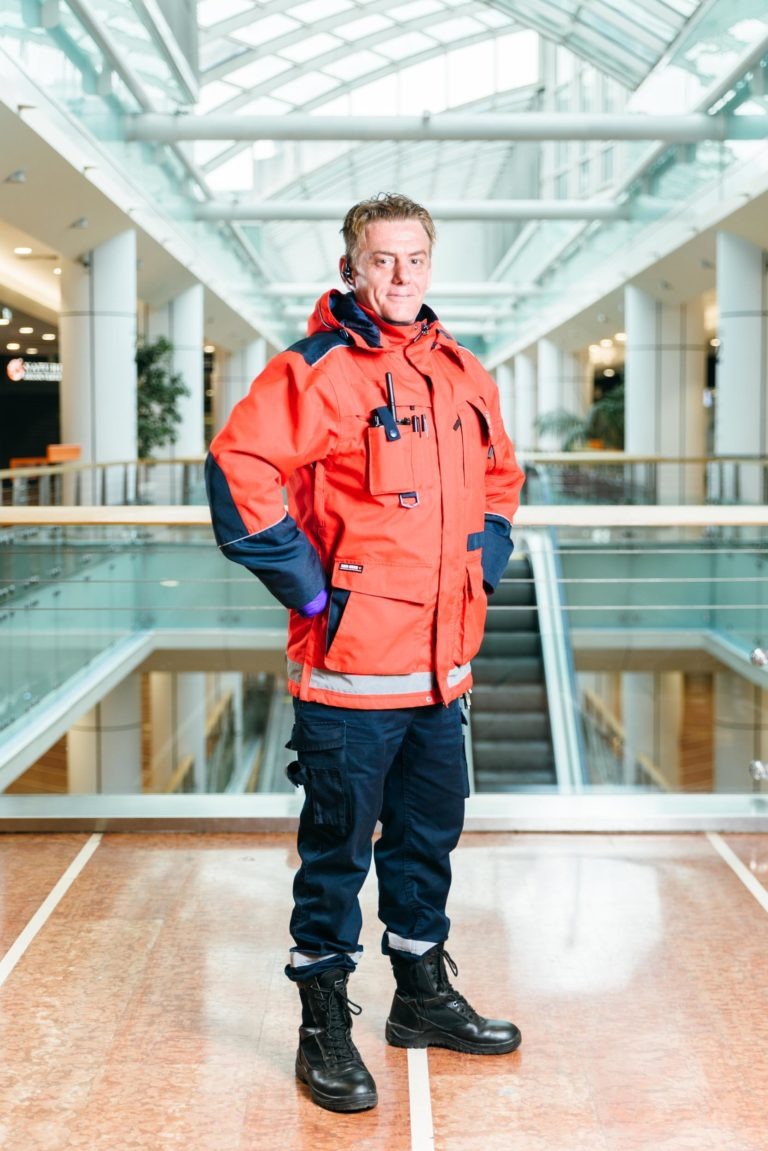 Un homme se tient debout, confiant, au centre du hall spacieux et moderne de l'Hyper-Héros du Centre Commercial Mériadeck, surmonté d'un plafond de verre. Il porte une veste rouge vif aux détails bleus, un pantalon bleu marine et de lourdes bottes noires. Les mains sur les hanches, il regarde directement la caméra. Des escaliers mécaniques et des rampes en verre sont derrière lui.