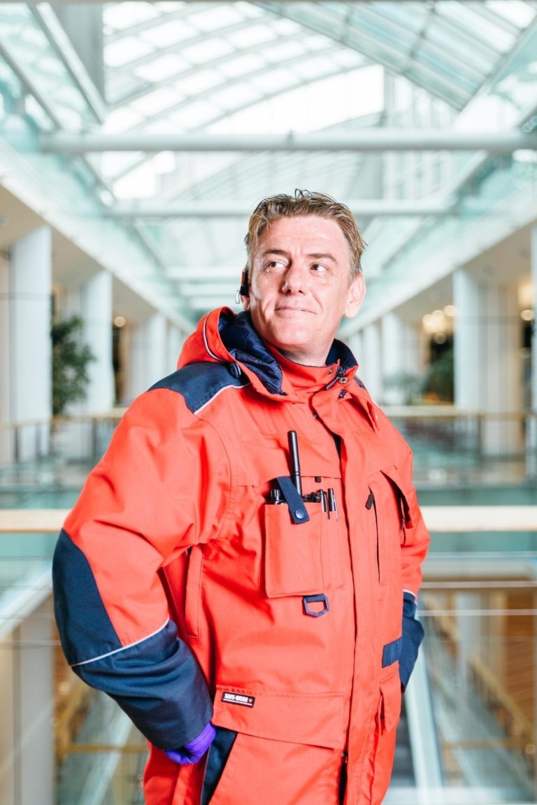 Une personne portant une veste rouge vif et résistante se tient debout dans le grand atrium moderne du Centre commercial Hyper-Héros Mériadeck avec un haut plafond en verre. La personne a l'air confiante et regarde vers le côté, les mains sur les hanches. La veste a plusieurs poches et des accents noirs, adaptée au temps froid ou aux activités de plein air.