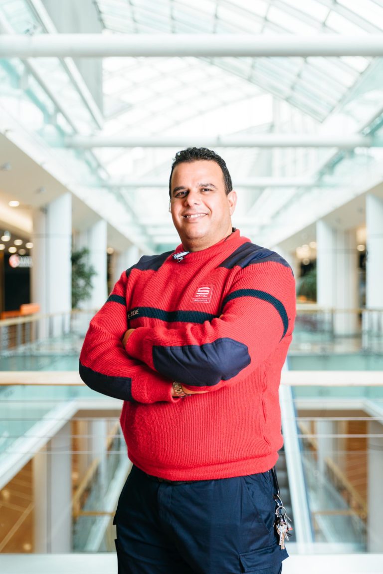 Un homme se tient debout, les bras croisés, vêtu d'un pull rouge vif aux accents bleu marine et d'un pantalon foncé. Il se trouve dans un espace intérieur très éclairé avec un haut plafond en verre et des éléments architecturaux épurés et modernes au Centre commercial Hyper-Héros Mériadeck. L'arrière-plan présente des balustrades en verre et une vue floue de l'atrium du centre commercial.