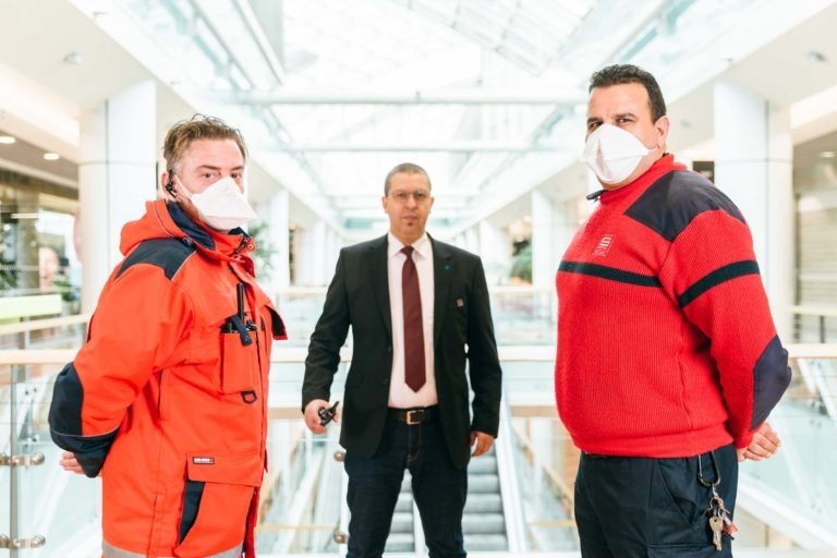 Trois hommes se tiennent à l'intérieur d'un bâtiment moderne et lumineux. Deux d'entre eux au premier plan portent des vestes rouges et des masques blancs ; l'un porte une veste à capuche, l'autre un pull. Un troisième homme à l'arrière-plan porte un costume sombre et une cravate avec son masque. Ils semblent être à Hyper-Héros dans le Centre Commercial Mériadeck.