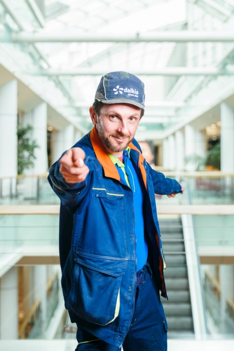 Une personne portant un uniforme de travail bleu et orange et une casquette bleue avec le mot « dalkia » pointe du doigt la caméra. Elle se trouve dans l'Hyper-Héros Centre Commercial Mériadeck, un espace intérieur moderne et bien éclairé avec de grandes fenêtres en verre et un escalator visible en arrière-plan. La personne a une expression confiante et engageante.