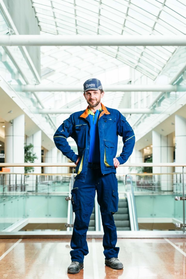 Un homme en uniforme de travail bleu et casquette se tient debout avec assurance dans le centre commercial Hyper-Héros Mériadeck, lumineux et moderne, avec un haut plafond de verre et des escaliers mécaniques en arrière-plan. Les mains sur les hanches et portant une barbe, il interagit directement avec la caméra dans ce cadre commercial spacieux.