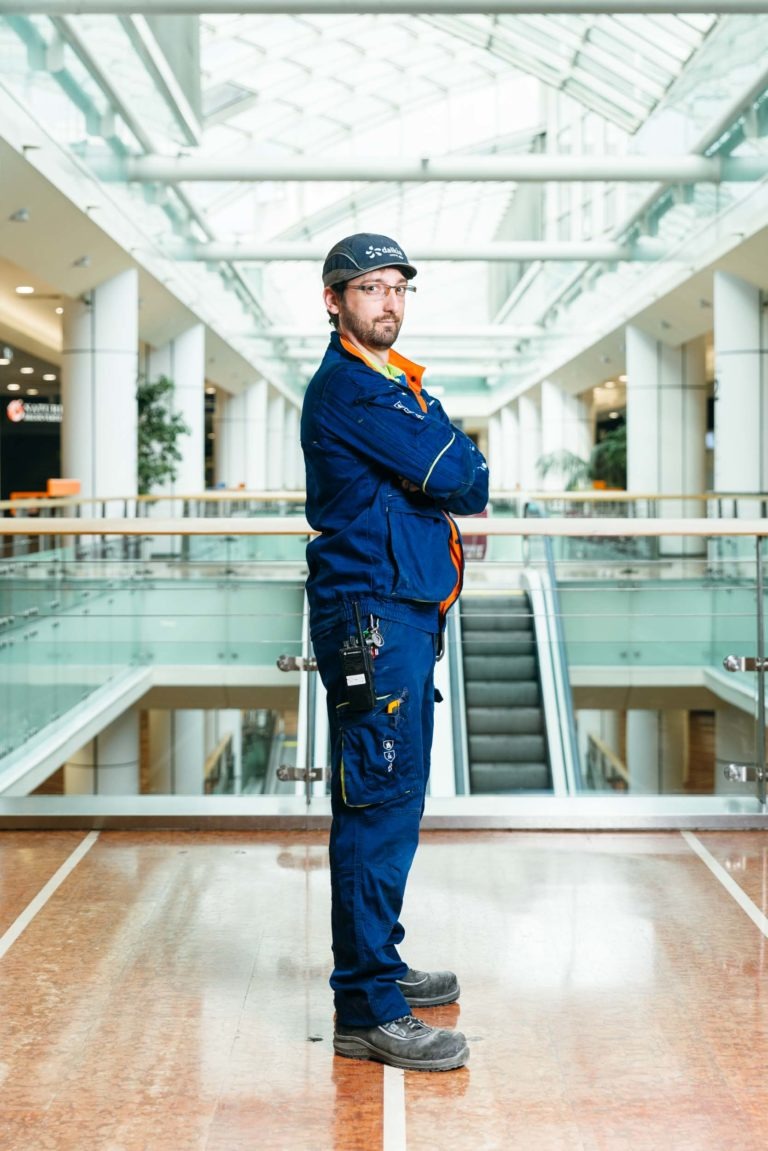 Un ouvrier du bâtiment se tient debout, les bras croisés, dans un atrium intérieur moderne et lumineux du Centre commercial Hyper-Héros Mériadeck. Il porte un uniforme bleu à bandes réfléchissantes, un casque de sécurité, des lunettes de sécurité, des gants et des bottes de travail. Un escalier mécanique et une rampe en verre sont visibles en arrière-plan.