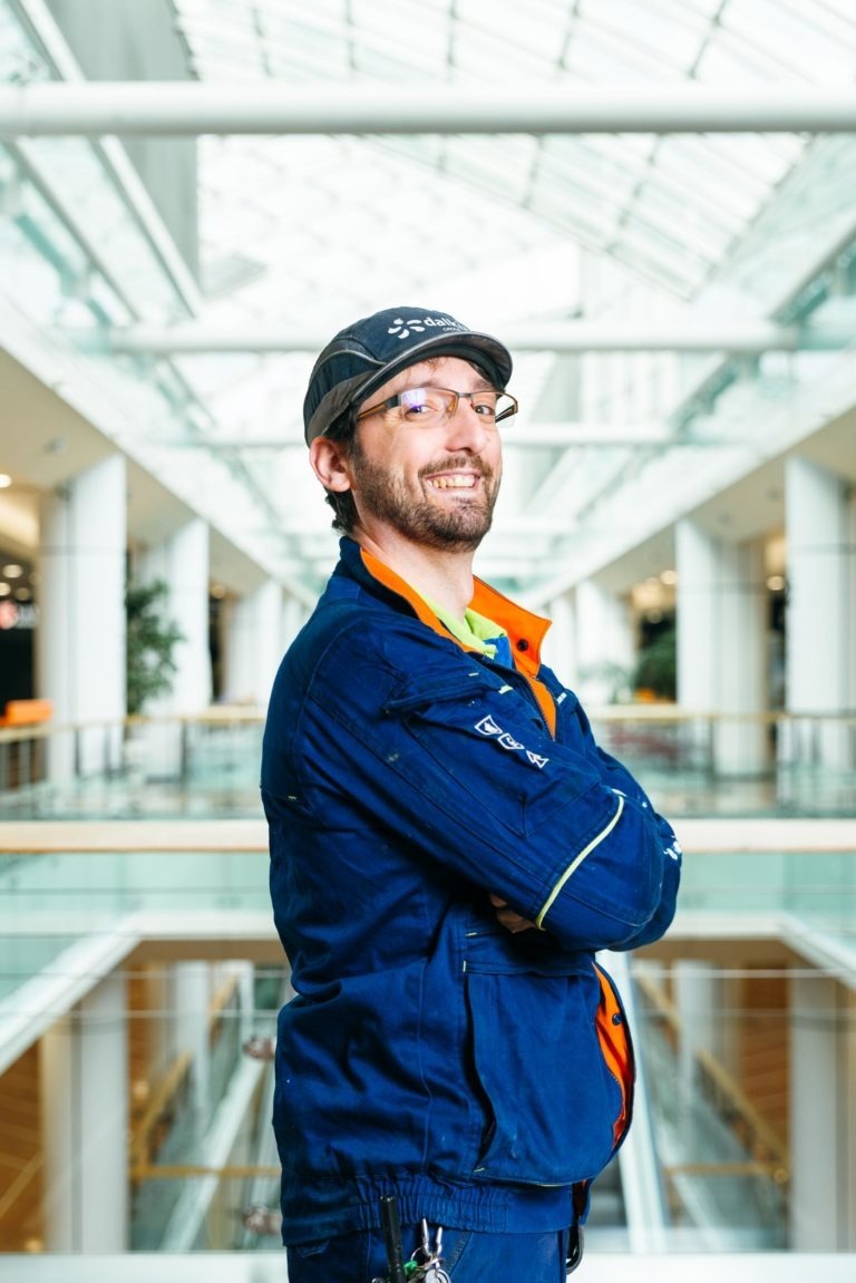 Un homme en uniforme de travail bleu et casquette se tient debout, les bras croisés, au Hyper-Héros Centre Commercial Mériadeck. Il porte des lunettes et une barbe, et il sourit. L'arrière-plan présente un intérieur moderne avec des balustrades en verre, de hautes colonnes blanches et un atrium éclairé par un puits de lumière.