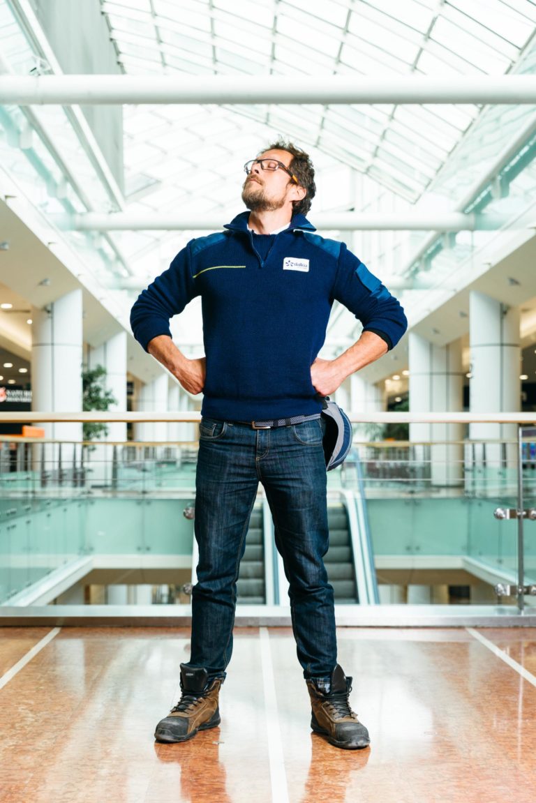 Un homme aux cheveux bouclés et aux lunettes se tient debout, les mains sur les hanches, à l'intérieur de l'Hyper-Héros Centre Commercial Mériadeck, un bâtiment moderne et lumineux avec des plafonds en verre. Il porte un pull bleu marine, un jean et des bottes de travail robustes, tenant un casque de sécurité à ses côtés. L'arrière-plan révèle un escalator et des rampes.