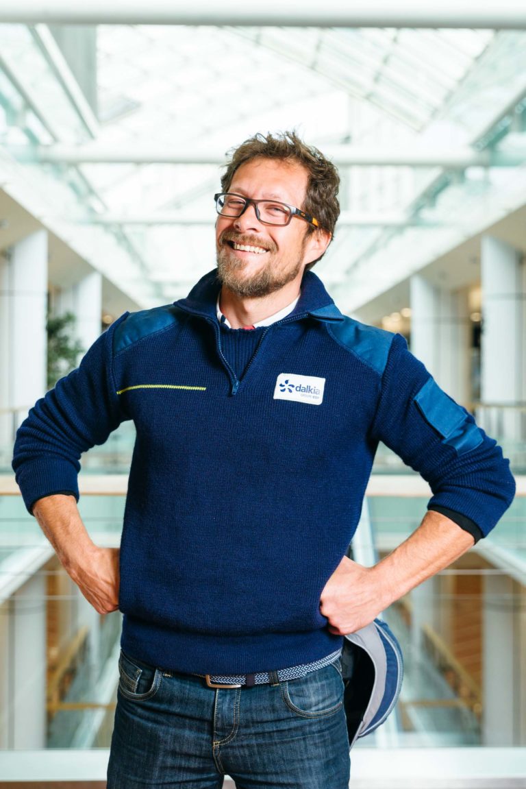 Un homme avec des lunettes, une barbe et un sourire amical se tient debout à l'intérieur, les mains sur les hanches. Il porte un pull bleu marine avec un logo Hyper-Héros et un jean foncé. L'arrière-plan présente l'atrium moderne au plafond de verre du Centre Commercial Mériadeck avec des éléments structurels blancs et une douce lumière naturelle qui y pénètre.