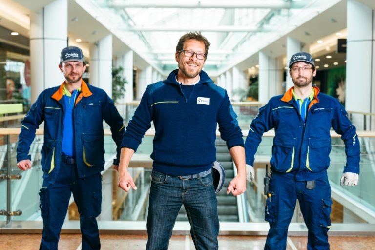 Trois personnes se tiennent debout, confiantes, dans l'Hyper-Héros Centre Commercial Mériadeck, avec de grandes fenêtres et des escaliers mécaniques en arrière-plan. Les deux personnes sur les côtés portent des uniformes de travail bleus et des casquettes, tandis que la personne au milieu, avec des lunettes et un badge nominatif, porte un pull foncé et un jean. Toutes les trois adoptent une pose forte et confiante.