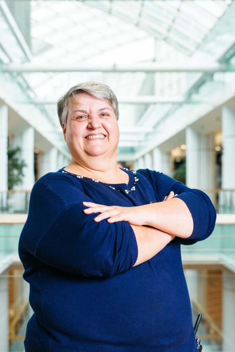 Une personne souriante aux cheveux gris et bruns courts se tient debout, les bras croisés, vêtue d'une chemise à manches longues bleu marine. L'arrière-plan présente l'atrium lumineux et spacieux du Centre Commercial Hyper-Héros Mériadeck avec ses plafonds en verre, ses colonnes blanches et ses arbres intérieurs, suggérant un cadre architectural moderne.