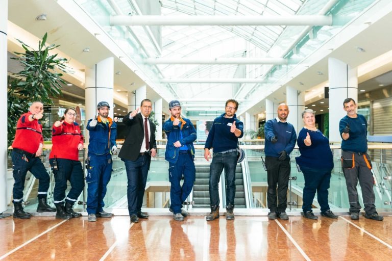 Un groupe de neuf personnes, vêtues d'un mélange d'uniformes de travail et de tenues décontractées, se tiennent à l'intérieur de l'Hyper-Héros du Centre Commercial Mériadeck, doté de plafonds en verre et de grandes fenêtres. Ils sont alignés côte à côte, chacun faisant un geste du pouce levé et souriant. Un escalator et de grandes plantes sont visibles en arrière-plan.