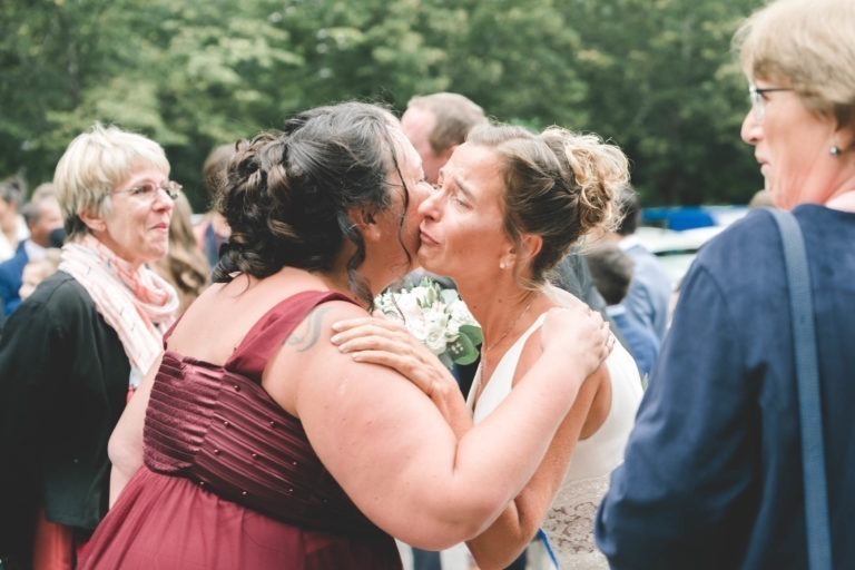 Deux femmes s'embrassent lors d'un événement en plein air. L'une porte une robe blanche, symbole de simplicité, tandis que l'autre porte une robe bordeaux. Elles se sourient chaleureusement au milieu d'autres participants en arrière-plan. La scène se déroule dans un espace vert et arboré où les gens discutent et discutent dans une atmosphère festive de générosité.