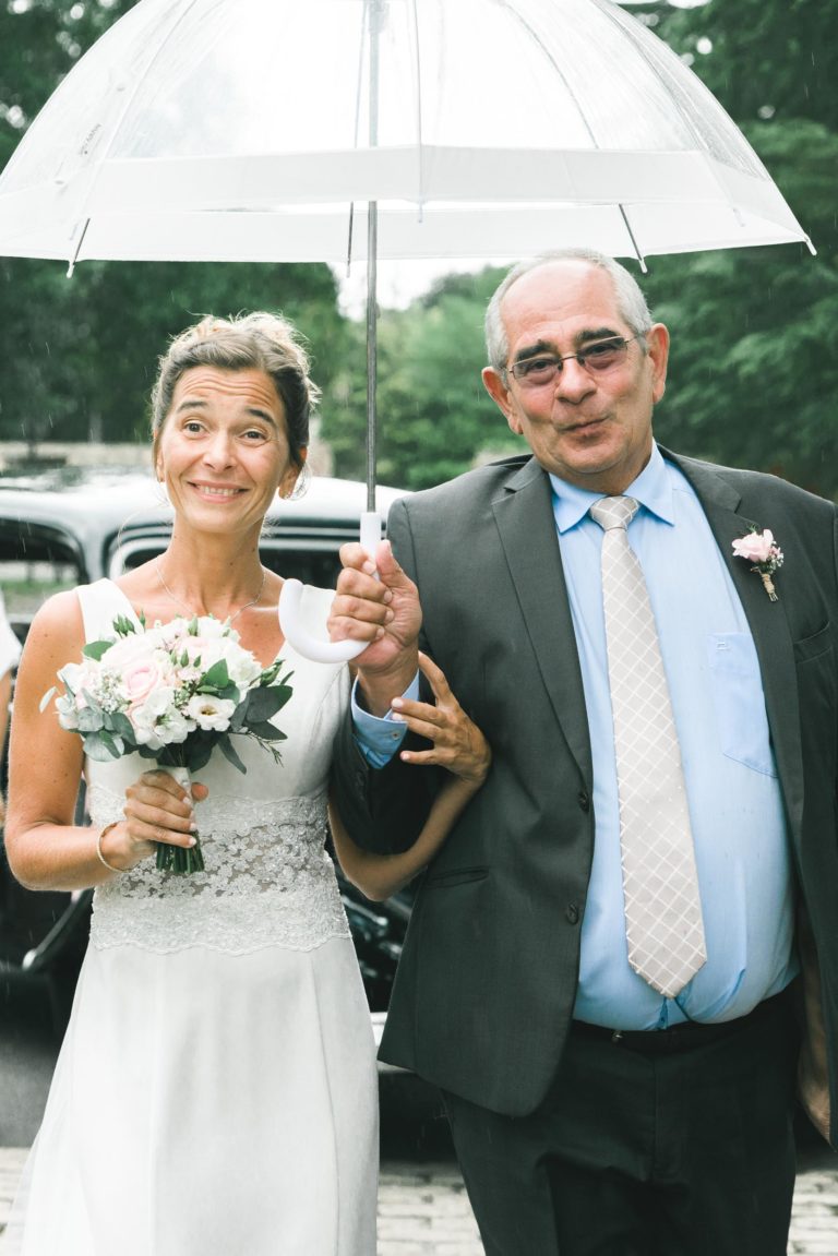 Une mariée en robe blanche et bouquet de fleurs sourit tout en tenant par le bras un homme plus âgé en costume gris et cravate. Partageant un parapluie transparent, ils se tiennent dehors avec des voitures anciennes et de la verdure en arrière-plan - une scène de simplicité joyeuse, peut-être après une cérémonie de mariage remplie de générosité.