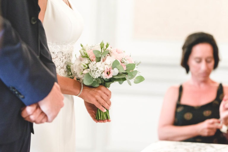 Une mariée tenant un bouquet de fleurs blanches et roses se tient debout à côté d'un marié vêtu d'un costume sombre. La robe blanche de la mariée, avec des détails en dentelle à la taille, respire la simplicité. À l'arrière-plan, une femme assise dans une robe noire à motifs floraux regarde vers le bas, partiellement floue.