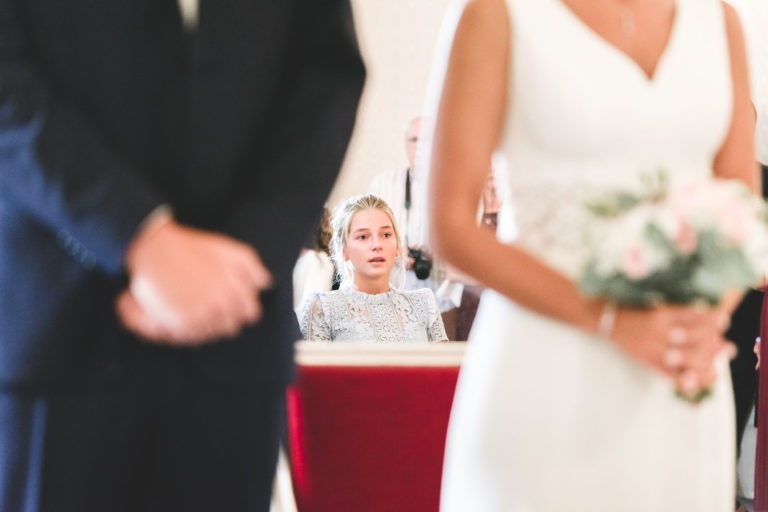 Dans un décor de mariage, un couple de mariés se tient au premier plan, légèrement flou. Le marié porte un costume sombre, la mariée porte une robe blanche simple et tient un bouquet. À l’arrière-plan, une femme assise et vêtue d’une robe bleu clair semble émue, son attention étant concentrée sur le mariage du couple.