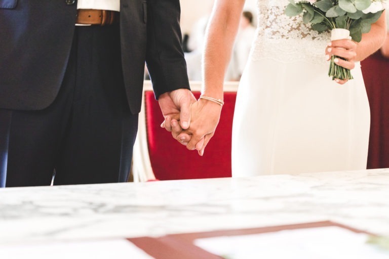 Un couple se tient main dans la main, vêtu d'une tenue formelle symbolisant la simplicité de leur lien. L'homme porte un costume noir et la femme une robe blanche avec des détails en dentelle sur le haut. Ils sont à côté d'une table en marbre avec des documents flous au premier plan, sur fond sombre et doucement éclairé.