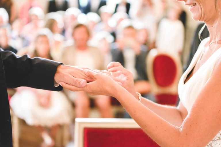 Gros plan d'une cérémonie de mariage où la mariée passe l'anneau au doigt du marié. Tous deux sont habillés de manière formelle ; la robe blanche de la mariée et le costume sombre de son époux sont partiellement visibles. L'arrière-plan montre un public assis sur des chaises rouges et beiges, floues, créant une atmosphère douce et intime, pleine de simplicité.