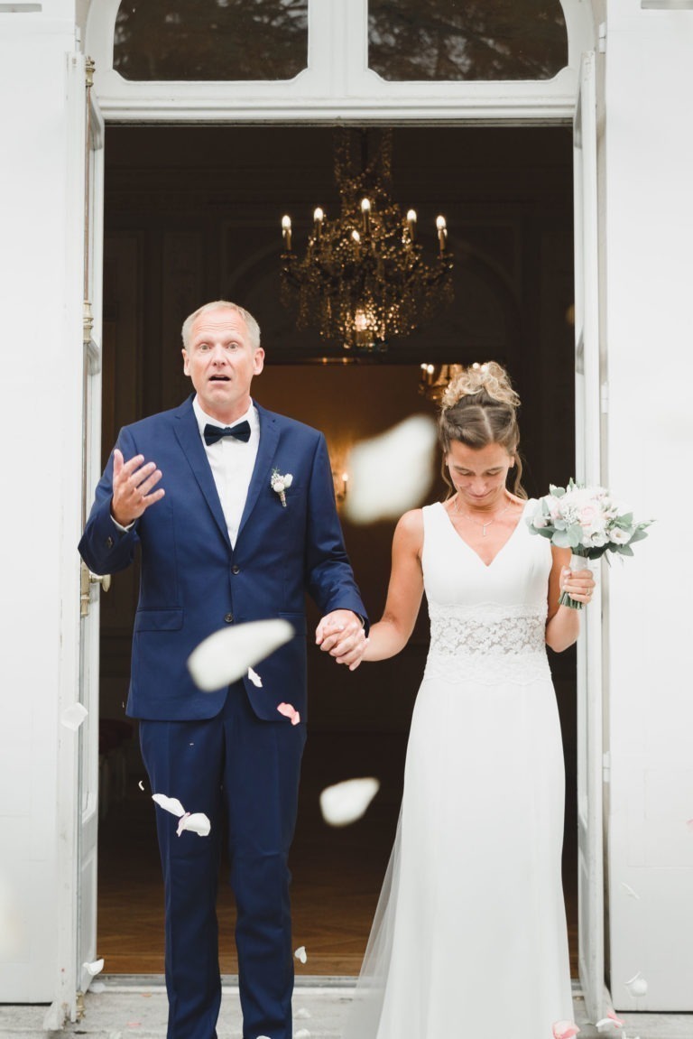 Un couple de jeunes mariés sort d'une salle de mariage par de grandes portes ouvertes. Le marié, en smoking bleu, a l'air surpris, tandis que la mariée, en robe blanche, tient un bouquet et regarde vers le bas avec un air de simplicité. Des pétales de roses blanches et roses flottent autour d'eux. Un lustre est suspendu à l'arrière-plan, symbolisant leur mariage.