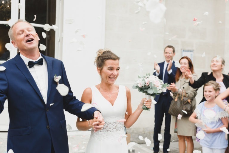 Un couple de mariés joyeux, se tenant la main, sourit tandis que des pétales de fleurs sont jetés sur eux. Le marié porte un costume bleu marine avec un nœud papillon, et la mariée porte une robe blanche, reflétant la simplicité, tout en tenant un bouquet. Derrière eux, les invités applaudissent et lancent des pétales, capturant l'atmosphère festive du jour de leur mariage.