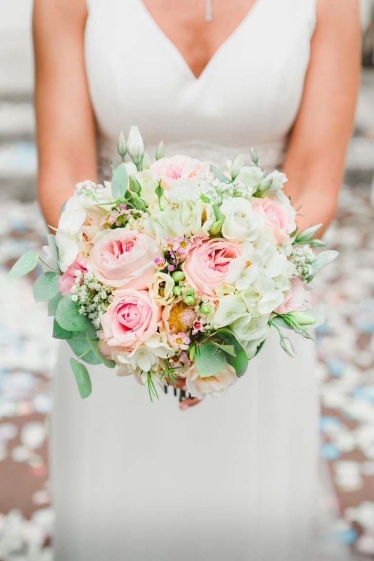 Une mariée vêtue d'une robe de mariée blanche tient un bouquet de fleurs à deux mains. Le bouquet, qui incarne la simplicité, comprend des roses roses, des hortensias blancs, des gypsophiles et du feuillage vert. L'arrière-plan est flou, avec des pétales de fleurs éparpillés. L'accent est mis sur les fleurs et le torse de la mariée.