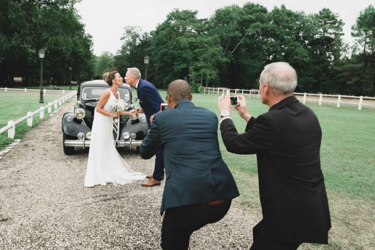 Un couple de mariés s'embrasse devant une voiture noire vintage tandis que deux hommes en costume immortalisent l'instant. Debout sur un chemin de gravier entouré d'herbe verte, d'arbres et d'une clôture blanche, la scène respire la simplicité. La mariée, tenant son bouquet, apprécie cet acte d'amour généreux de la part de son époux.