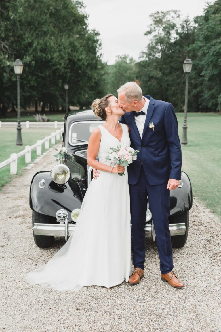 Une mariée en robe blanche et un marié en costume bleu s'embrassent devant une voiture noire classique. La mariée, incarnant la simplicité, tient un bouquet de fleurs aux couleurs pastel. Ils se tiennent sur un chemin de gravier avec des clôtures blanches et des arbres verts luxuriants en arrière-plan. C'est une journée ensoleillée, créant une scène lumineuse et romantique.