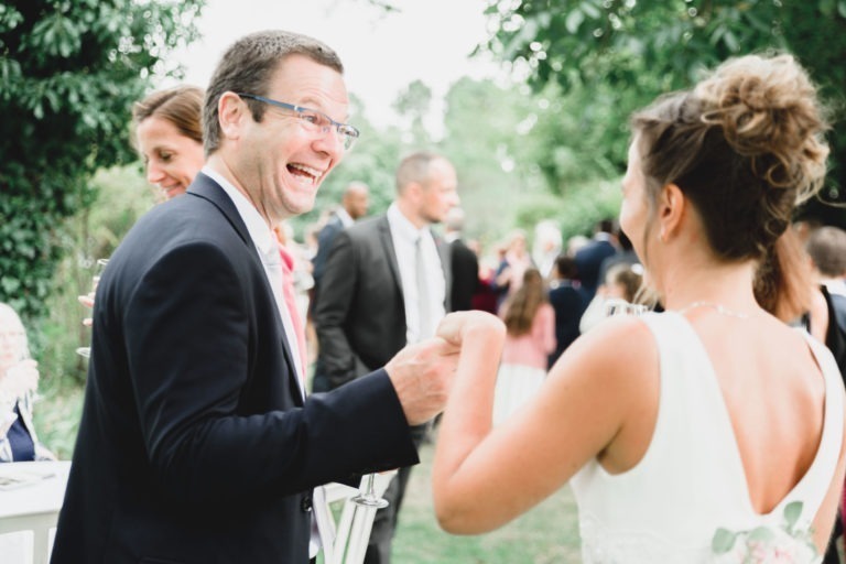 Un homme portant des lunettes et un costume sombre discute joyeusement avec une femme en robe blanche, tous deux souriants et se tenant la main. Ils se tiennent au milieu d'un groupe de personnes en tenue de soirée lors d'un événement en plein air, entouré de verdure luxuriante. L'atmosphère est festive et animée, suggérant une célébration de la simplicité et de la générosité.