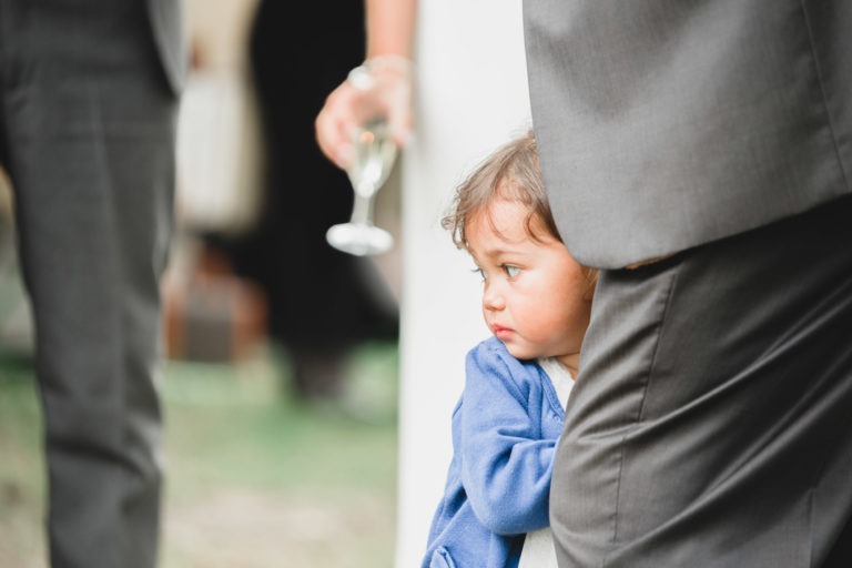 Un jeune enfant aux cheveux bruns, vêtu d'une veste bleue, apparaît derrière la jambe d'un adulte vêtu d'un costume gris. L'enfant a l'air légèrement inquiet. L'adulte tient un verre de vin dans une main, incarnant un air de simplicité. L'arrière-plan est flou, mettant en valeur la scène au premier plan.