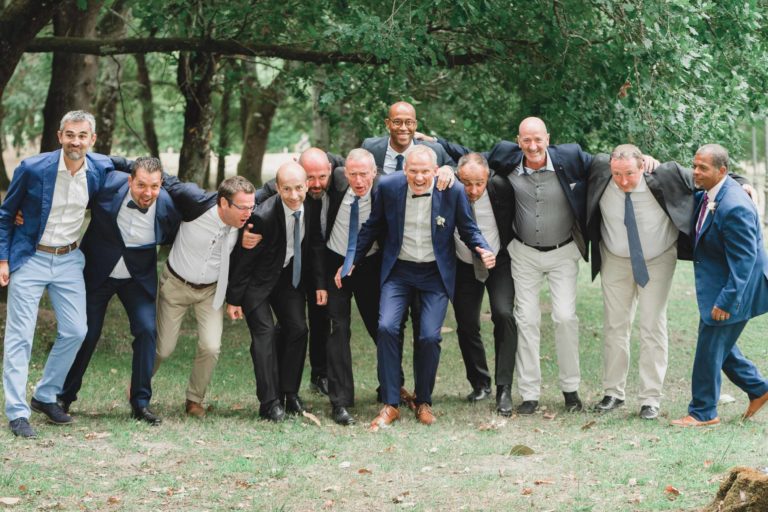 Onze hommes en tenue de soirée se tiennent côte à côte dans un décor extérieur, souriant et se tenant les uns les autres, les bras autour des épaules. Le décor comprend des arbres verts luxuriants et une zone herbeuse, créant une atmosphère animée et festive de simplicité et de générosité.