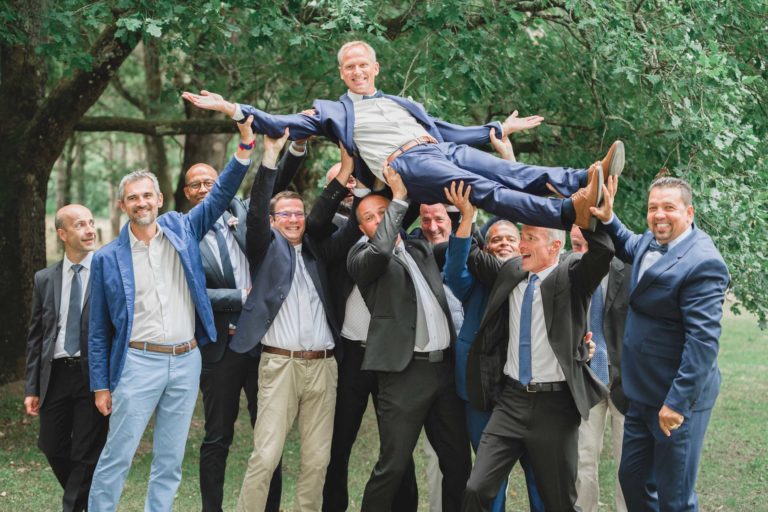 Un groupe de onze hommes en costume et cravate se trouve à l'extérieur, avec de la verdure en arrière-plan. Ils sourient et soulèvent horizontalement dans les airs l'un des hommes, qui porte un costume bleu marine, tandis qu'il écarte les bras et les jambes dans une pose joyeuse. L'atmosphère est festive et joyeuse, incarnant la simplicité de la véritable camaraderie.