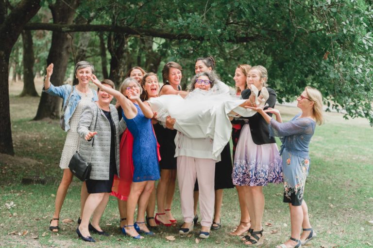 Un groupe de douze femmes se trouve à l'extérieur, réunies dans un parc avec des arbres verts luxuriants en arrière-plan. Elles sourient et font la fête en tenant une autre femme vêtue d'une robe de mariée blanche au-dessus de leurs épaules. L'ambiance est joyeuse et festive, incarnant la simplicité et la générosité du mariage ; tout le monde est habillé de tenues colorées et semi-formelles.