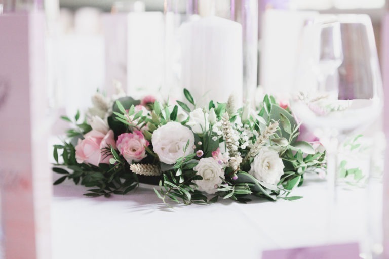 Gros plan d'une composition florale sur une table. La composition, symbole de simplicité, comprend des fleurs blanches, roses et roses accentuées de verdure. Autour de la pièce maîtresse se trouvent des éléments de vaisselle flous, notamment un verre à vin à droite. Le décor dégage une ambiance élégante et festive.