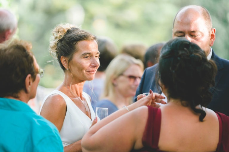 Une femme en robe blanche tient un verre et discute avec un groupe de personnes lors d'un événement en plein air. Le groupe comprend un homme en costume et une femme en robe bordeaux. Les personnes à l'arrière-plan sont habillées de manière décontractée et la scène respire la simplicité et l'élégance décontractée au milieu de la verdure et d'un éclairage doux.