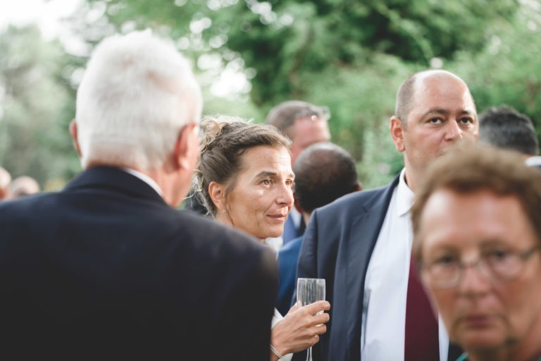 Un groupe de personnes est réuni à l'extérieur pour discuter. Une femme au centre tient un verre et discute avec simplicité. Plusieurs hommes sont à proximité, habillés en tenue de soirée. L'arrière-plan présente de la verdure, suggérant que l'événement se déroule dans un jardin ou un parc.