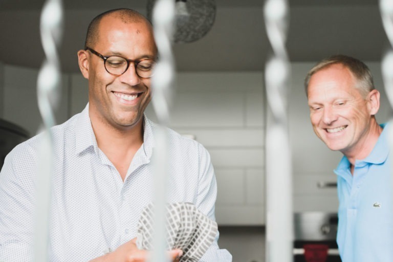 Deux hommes dans une cuisine sourient en travaillant. L'homme de gauche, portant des lunettes et une chemise claire, essuie un plat avec une serviette à carreaux. L'homme de droite, vêtu d'un polo bleu clair, regarde avec bonheur. La photo est prise à travers des barres floues au premier plan, capturant leur simplicité et leur chaleur.