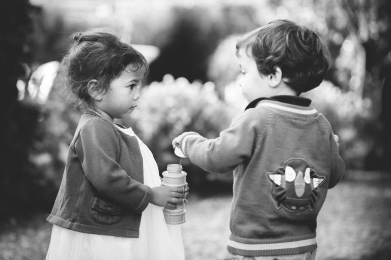 Une photo en noir et blanc de deux jeunes enfants en plein air. La fille à gauche tient une baguette à bulles et regarde le garçon à droite, qui fait des gestes de la main. Tous deux sont habillés avec simplicité et désinvolture, le garçon portant une veste avec un personnage de dessin animé dans le dos. L'arrière-plan est flou.