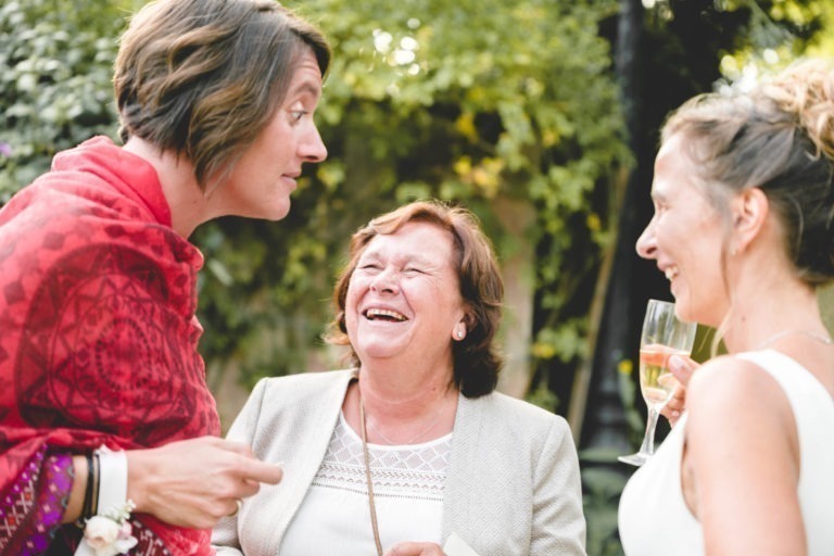 Trois femmes discutent joyeusement en plein air. La femme de gauche, vêtue d'un châle rouge, interagit avec animation. La femme du milieu rit ouvertement, vêtue d'un blazer clair. La femme de droite, dans une tenue blanche qui respire la simplicité, tient un verre de champagne et sourit. Un feuillage vert est visible à l'arrière-plan.