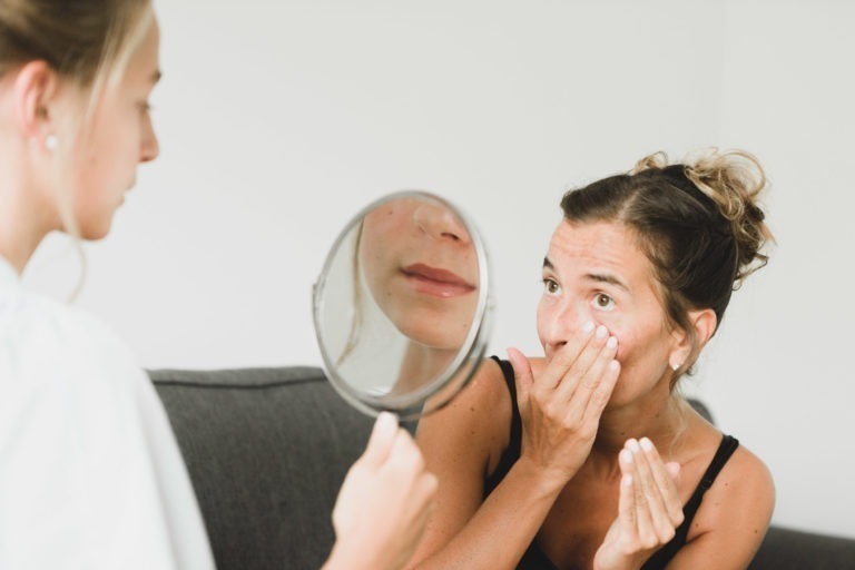 Une femme, les cheveux en chignon, se touche le visage sous l'œil, l'air inquiet. Une autre personne, dont le visage est en partie visible, lui tend un miroir rond, reflétant un gros plan de ses lèvres. Elles se trouvent dans une pièce bien éclairée avec un arrière-plan simple et clair.