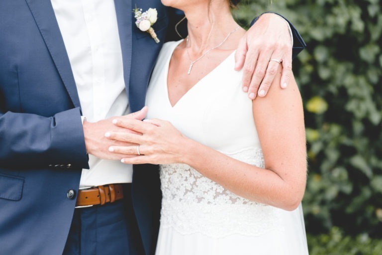 Un couple en tenue de mariage s'embrasse devant un fond de feuilles vertes. La mariée porte une robe blanche avec des détails en dentelle, qui respire la simplicité, et le marié est vêtu d'un costume bleu marine et d'une chemise blanche. Tous deux ont des alliances visibles sur leurs mains, le bras du marié enroulé autour de la mariée, symbolisant leur amour généreux.