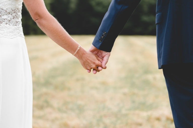 Une photo en gros plan d'un couple se tenant la main en plein air illustre la simplicité du véritable amour. La personne à gauche porte une robe blanche avec des détails en dentelle, tandis que la personne à droite porte un costume bleu foncé. L'arrière-plan présente un champ herbeux et une verdure floue, suggérant un cadre naturel.