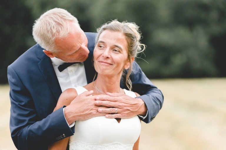 Un marié en costume bleu marine et nœud papillon noir enlace amoureusement sa future épouse par derrière. La mariée, vêtue d'une robe de mariée blanche, sourit avec contentement et regarde devant elle. Le couple se tient debout dans un décor extérieur avec un arrière-plan vert flou, incarnant la simplicité et la générosité de leur mariage.