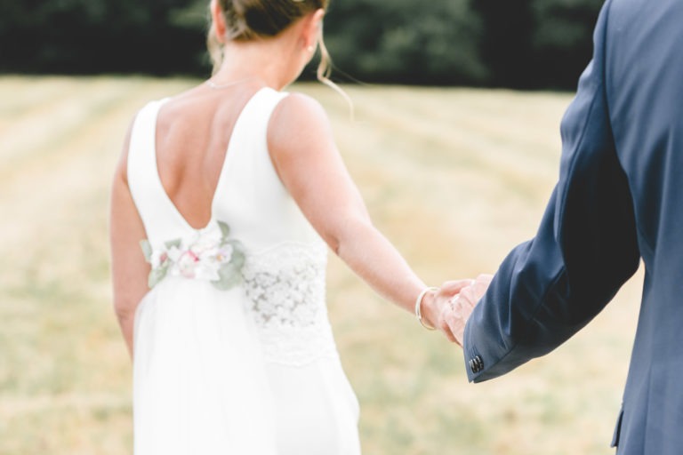 Une mariée dans une robe blanche sans manches avec des détails floraux à la taille tient la main d'une personne en costume sombre. Elles s'éloignent de la caméra à travers un champ, avec des arbres verts flous en arrière-plan. Les cheveux de la mariée sont coiffés en chignon. L'image respire la générosité et la simplicité de leur journée spéciale.