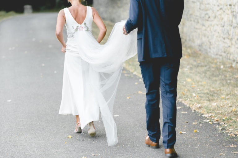 Une mariée en robe blanche marche aux côtés d'un homme en costume bleu marine et chaussures marron. Il tient une partie de son long voile flottant tandis qu'ils déambulent sur un chemin pavé bordé de verdure et d'un mur de pierre. Ils tournent le dos à l'objectif, capturant la simplicité et la générosité de leur mariage.