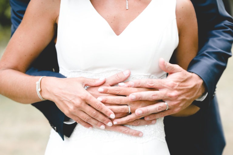 Gros plan sur un couple lors de leur mariage. Le marié, vêtu d'un costume sombre, enlace doucement la mariée, qui porte une robe blanche aux détails en dentelle. Les deux ont les mains entrelacées sur la taille de la mariée, mettant en valeur leurs alliances. La mariée porte un bracelet et un collier délicats, incarnant la simplicité.