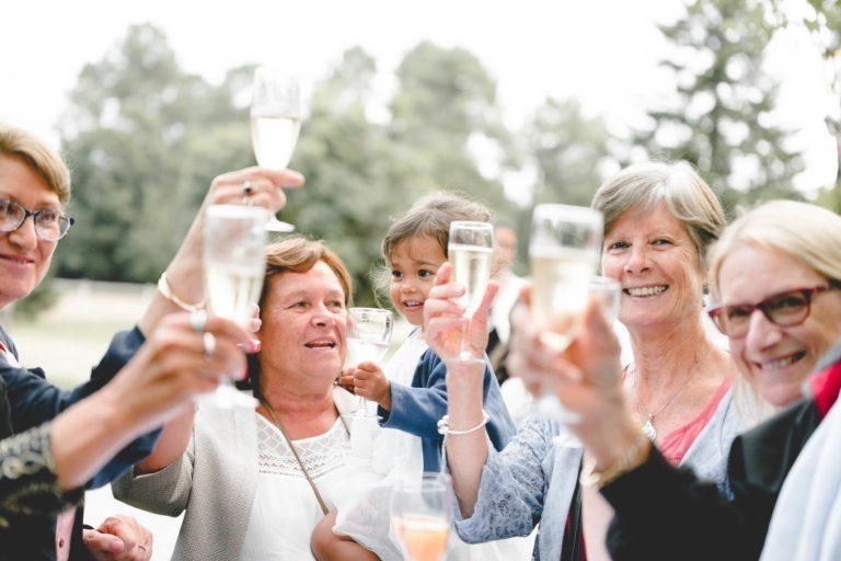 Un groupe de cinq personnes lèvent des coupes de champagne pour célébrer la fête en plein air. L'une des personnes tient dans ses bras un petit enfant qui tient également une coupe. L'arrière-plan est composé de verdure et d'arbres, suggérant un parc ou un jardin. La scène incarne la simplicité et la joie du moment, tout le monde paraissant heureux et joyeux.