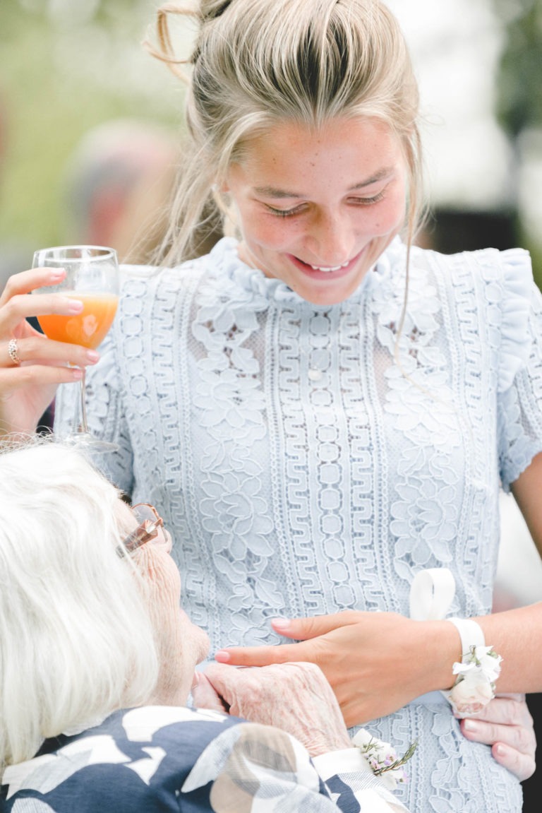 Une jeune femme vêtue d'une délicate robe en dentelle bleu clair tient un verre contenant une boisson orange et sourit à une femme âgée aux cheveux blancs. La femme âgée, vêtue de vêtements à motifs sombres, tient et embrasse la main de la jeune femme avec une générosité sincère, incarnant la simplicité de leur lien. Le décor semble être celui d'un rassemblement en plein air.