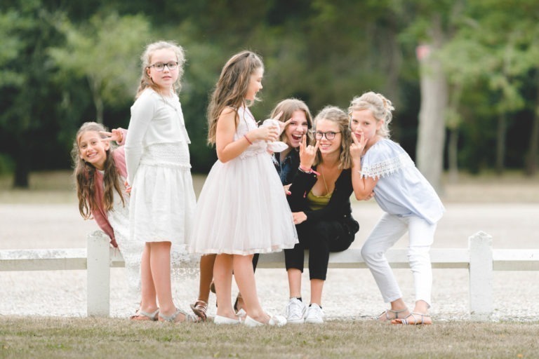 Un groupe de sept enfants et adolescents sont réunis à l'extérieur, près d'une clôture blanche. Quatre filles se tiennent debout, certaines portant des robes et des vêtements décontractés, tandis que trois enfants se penchent vers l'appareil photo et posent joyeusement. Les arbres et la verdure sur l'arrière-plan flou ajoutent à la simplicité de la scène, suggérant un décor de parc.