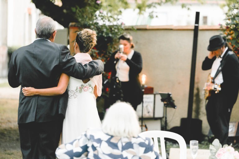 Un homme âgé et une mariée, vus de dos, s'embrassent lors d'une réception de mariage en plein air. En arrière-plan, une femme parle dans un micro tandis qu'un musicien portant un chapeau joue de la guitare. Une personne aux cheveux blancs est assise sur une chaise blanche au premier plan, observant le couple avec simplicité et chaleur.