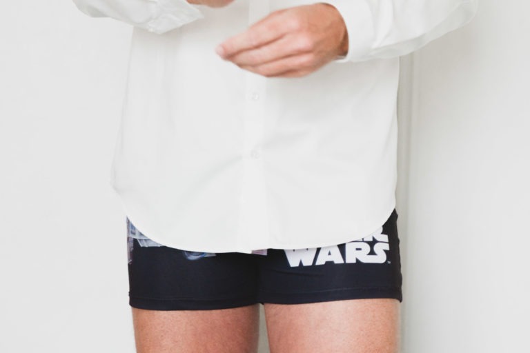 Une personne est partiellement visible du cou jusqu'à mi-cuisse, portant une chemise blanche et un caleçon noir à thème Star Wars. Les manches de sa chemise sont légèrement retroussées, incarnant un sentiment de simplicité. Les bras de l'individu sont légèrement levés, ajustant quelque chose hors du cadre. L'arrière-plan est uni et blanc.