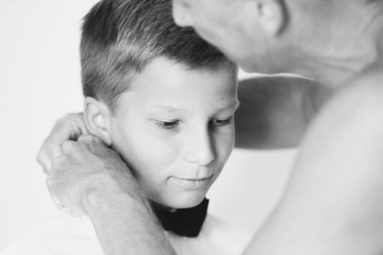 Gros plan en noir et blanc d'un jeune garçon en chemise blanche et nœud papillon noir qui reçoit l'aide d'un adulte pour ajuster ses vêtements. Le garçon baisse les yeux avec un léger sourire tandis que l'adulte, les mains près du cou du garçon, se concentre sur la tâche. La simplicité de leurs gestes souligne un moment d'intimité et de générosité.